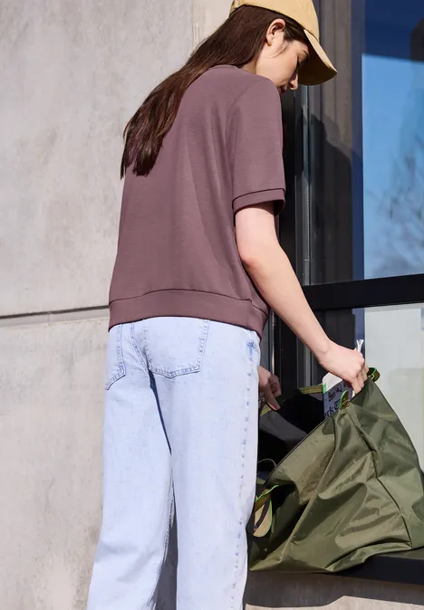 Frau mit lila Poloshirt, heller Jeans, beiger Kappe hält grüne Tasche vor moderner Fassade.